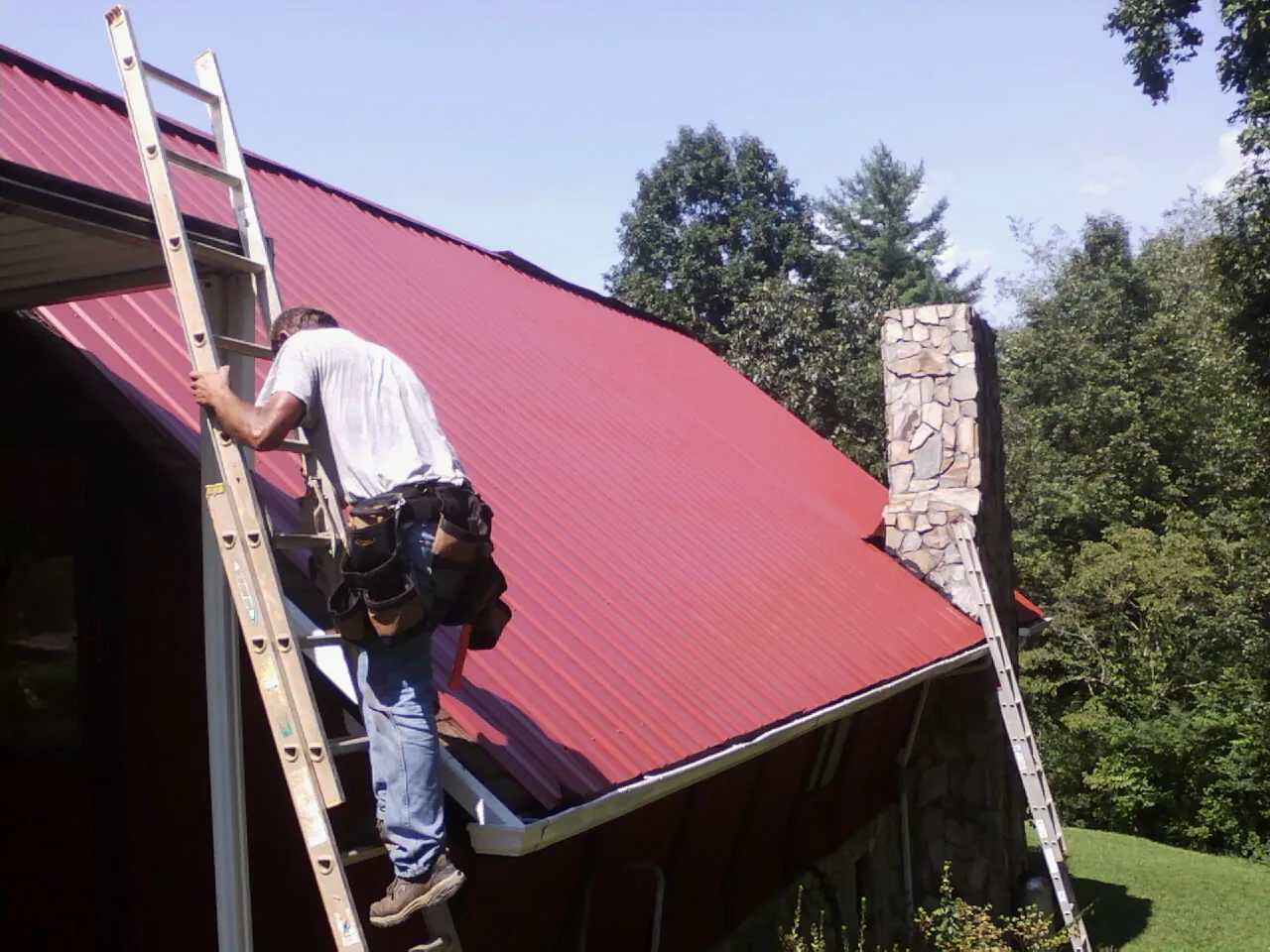 Roofer on a ladder beside a red metal roof and stone chimney — Soffit & Fascia Repair by Roofing Brothers in Dry Ridge